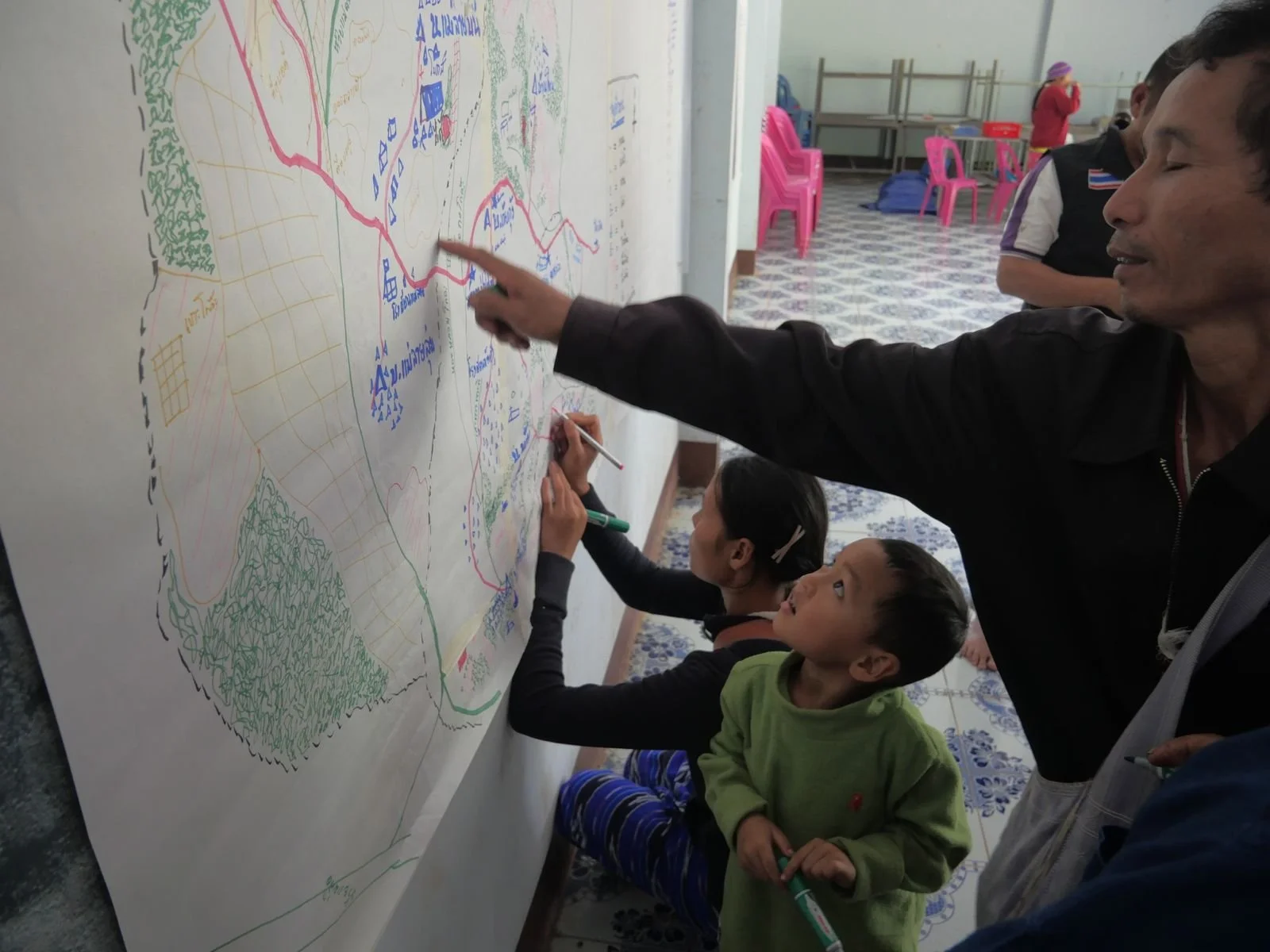People, including children and an adult man, drawing and explaining a colorful map or diagram on a large whiteboard or paper, in a room with tiled floor and pink plastic chairs in the background.