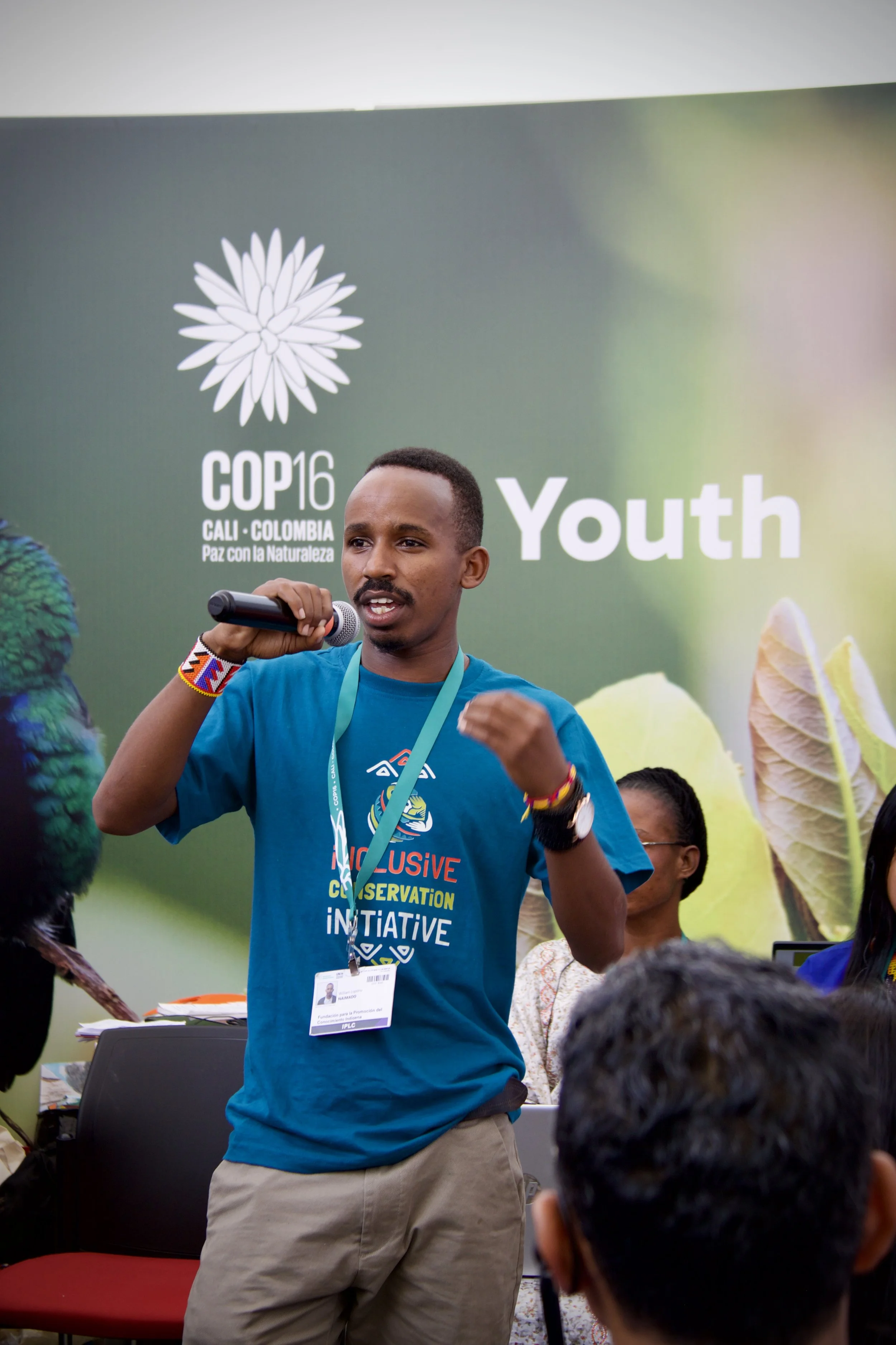 A young Indigenous Kenyan man speaking into a microphone at a conference or event with a green backdrop that reads 'COP16 Cali Colombia Paz con la Naturaleza Youth.'