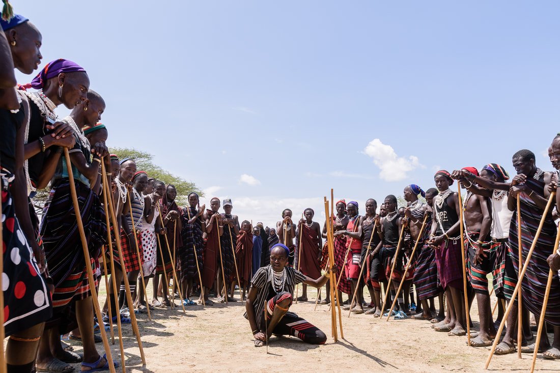 Group of Maasai people standing in a circle outdoors, with a woman in traditional attire kneeling in the center, holding a staff, under a clear blue sky.