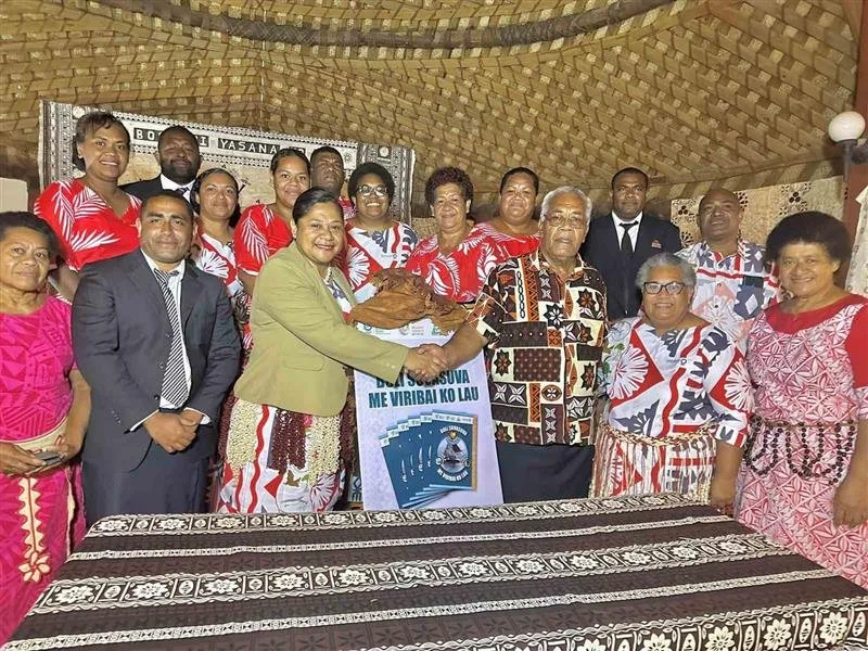 Group of people standing inside a decorated room, some wearing colorful traditional clothing, celebrating and shaking hands around a large poster in the Pacific.