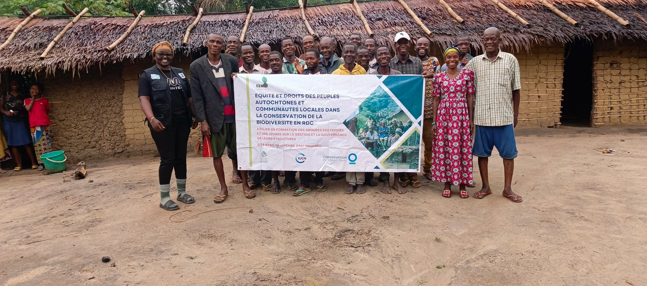 Group of people holding a banner in front of traditional mud-brick building with a thatched roof in a rural setting in the DRC.
