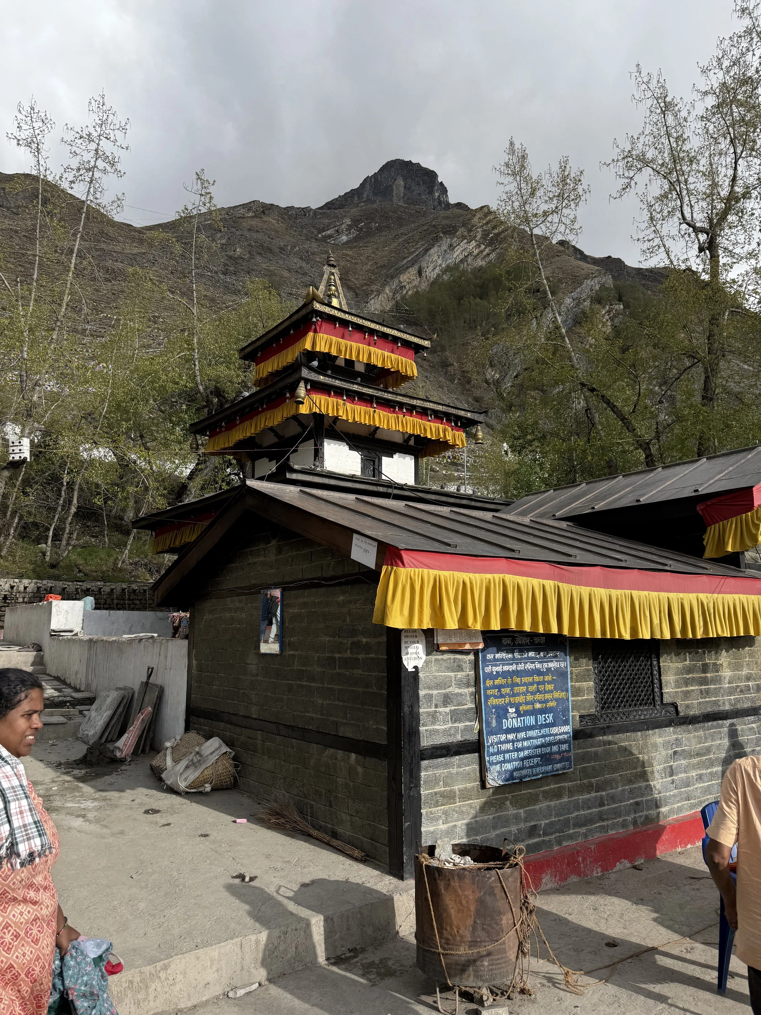 A small temple with multiple tiers, decorated with red and yellow drapes, situated at the base of a mountain. There are people nearby and a donation desk outside in Nepal.
