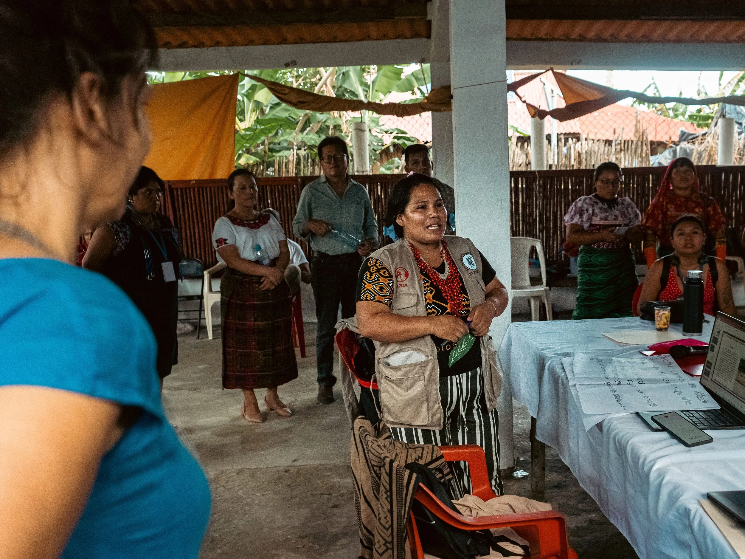 A group of Indigenous People gathered inside a semi-open space, with some standing and some seated around a table. A woman in the center wears a beige vest and speaks to the group, with others listening attentively in Peru in the Southwest Amazon.
