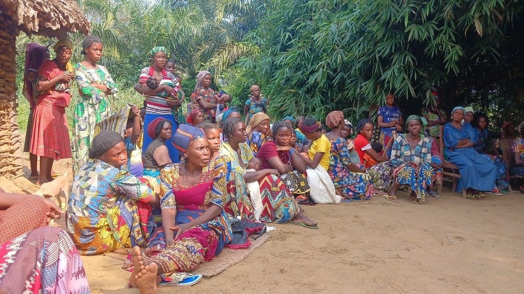 Group of women and children sitting and standing outdoors in a lush, green setting with large leafy plants in the background in the DRC.