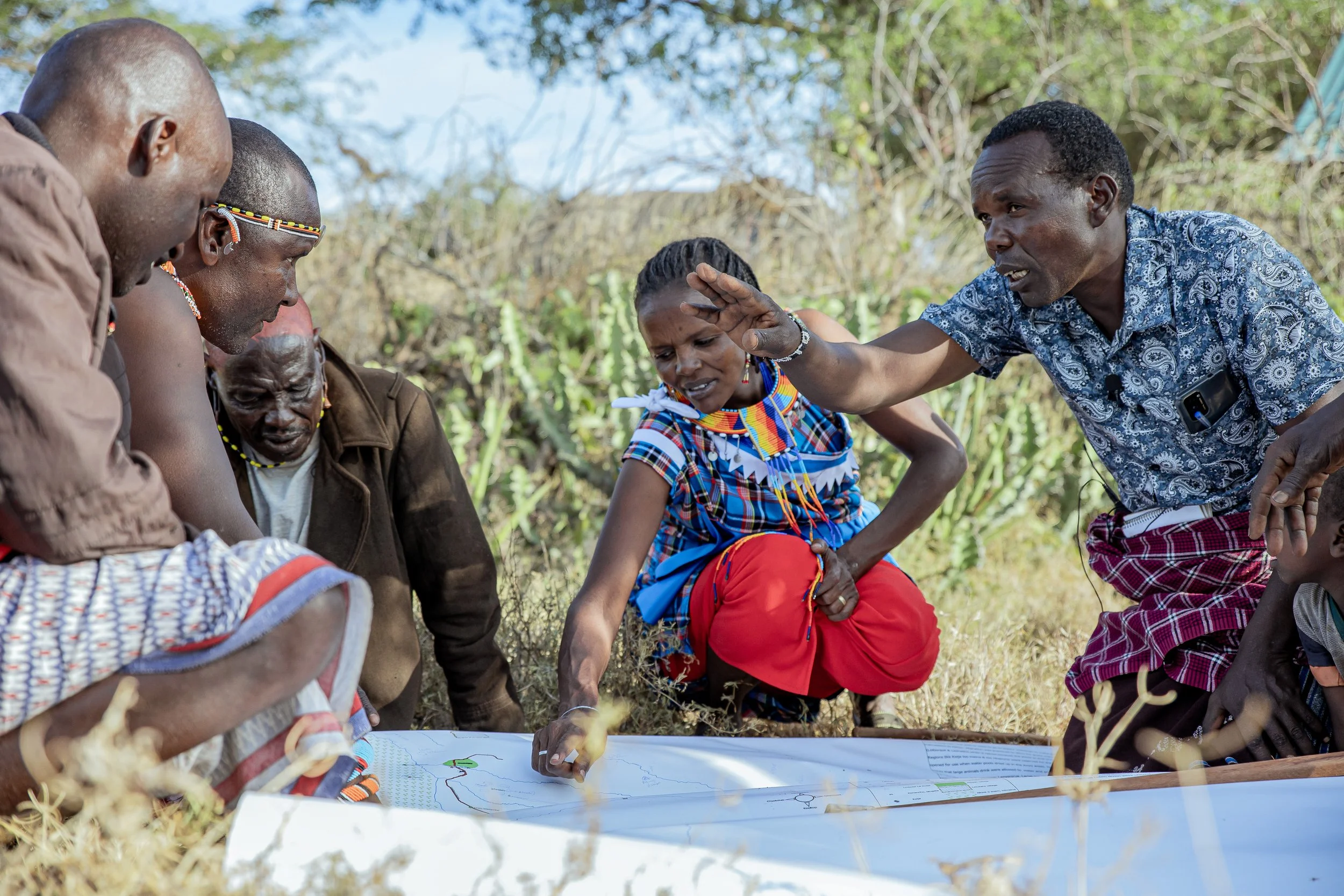 A group of people in Kenya, including a woman and a man with a phone in his pocket, gather around a large map or diagram outdoors, with some pointing and discussing, surrounded by bushes and trees in a rural setting.