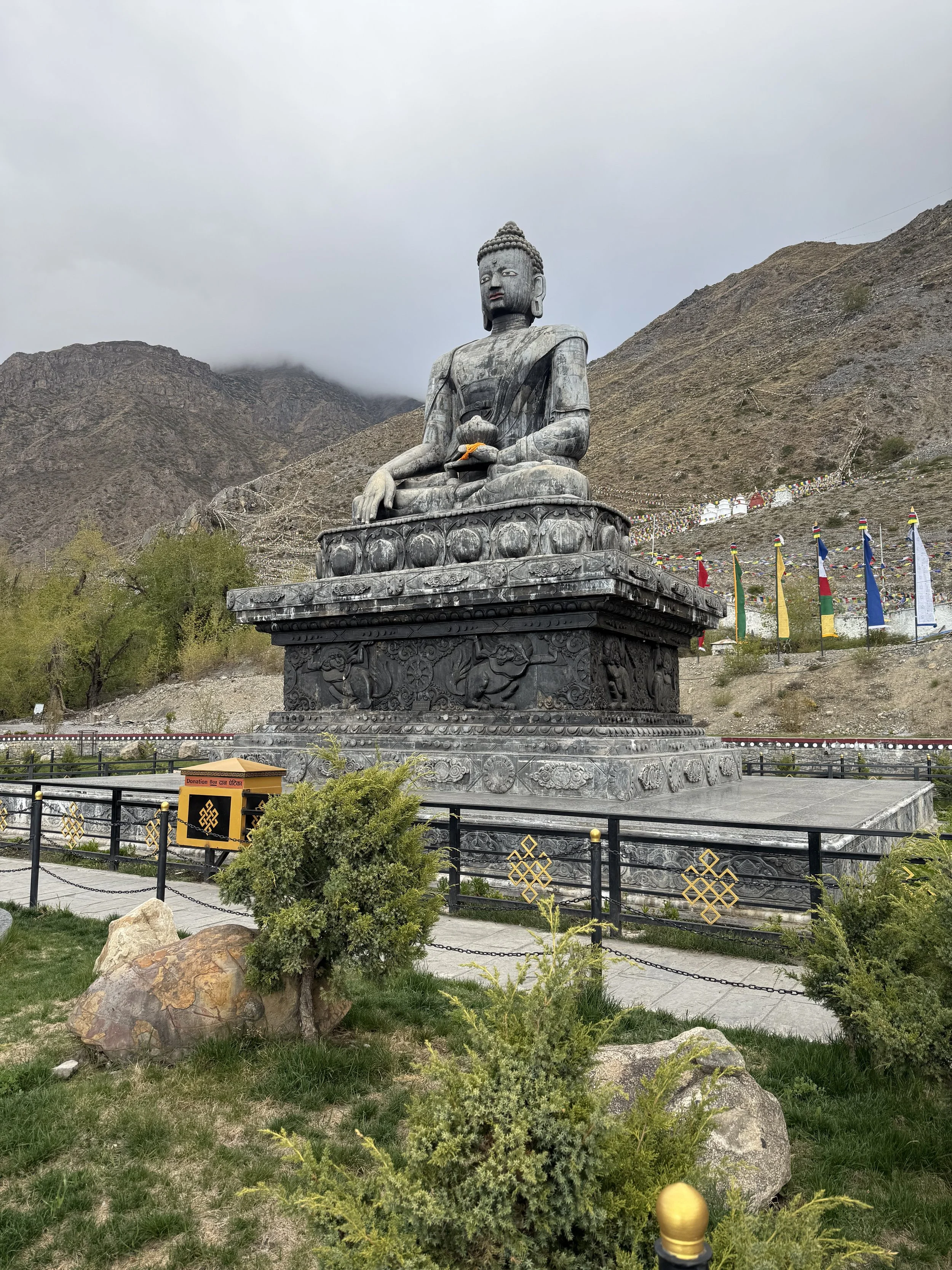 Large black stone statue of Buddha sitting in a meditative pose, situated outdoors against a mountainous background with cloudy skies and colorful prayer flags in Nepal.