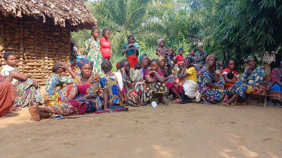 A group of women and children sitting and standing outdoors in a rural village, with traditional thatched huts and lush green trees in the background.