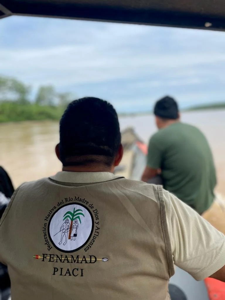 Indigenous People in a boat, with the back of a person wearing a beige shirt with a logo and text that reads 'FENAMAD PLACI', overlooking a river with trees on the far bank in the Southwest Amazon in Peru.