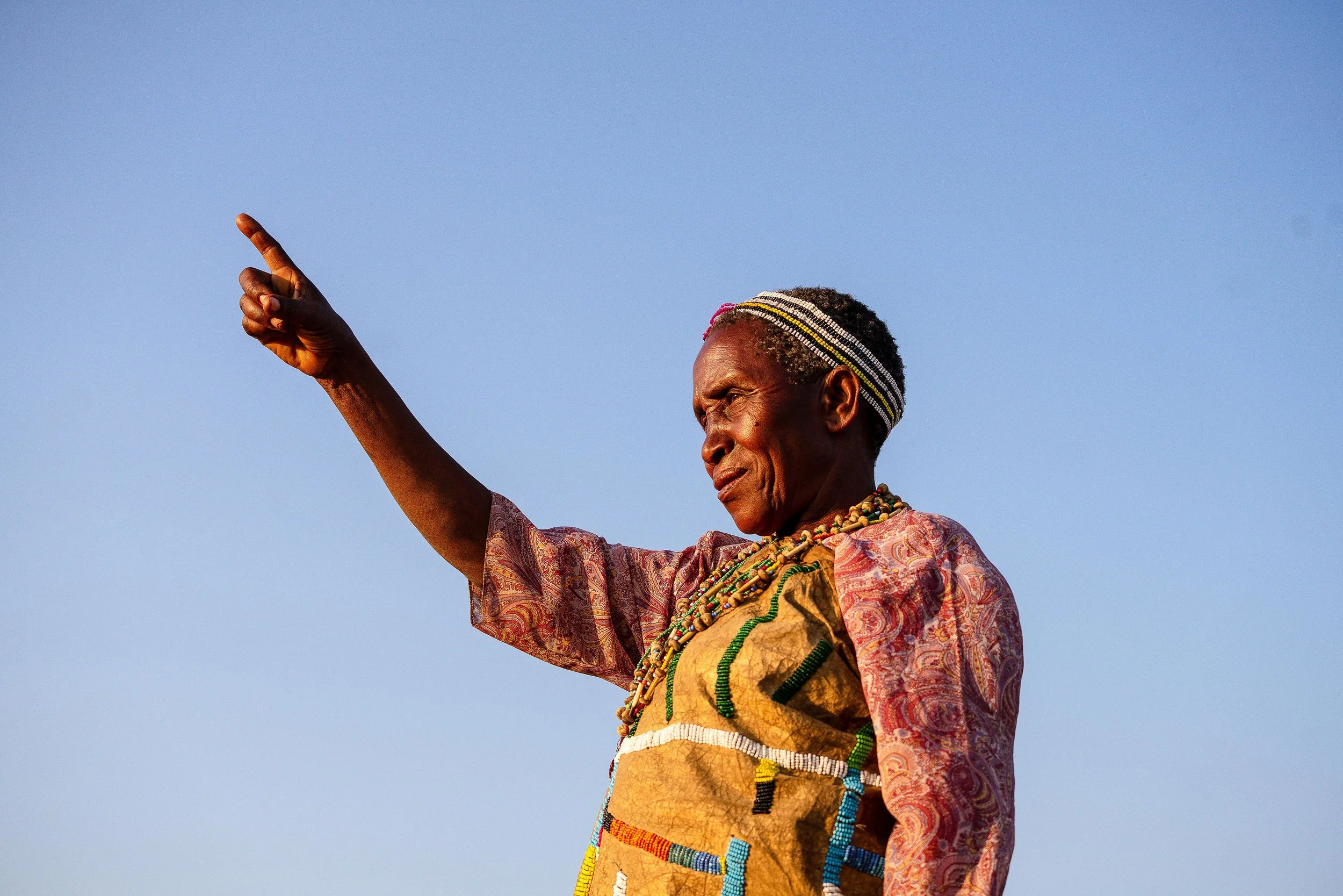 An older Indigenous Tanzanian woman dressed in traditional attire with colorful necklaces, pointing into the distance against a clear blue sky.