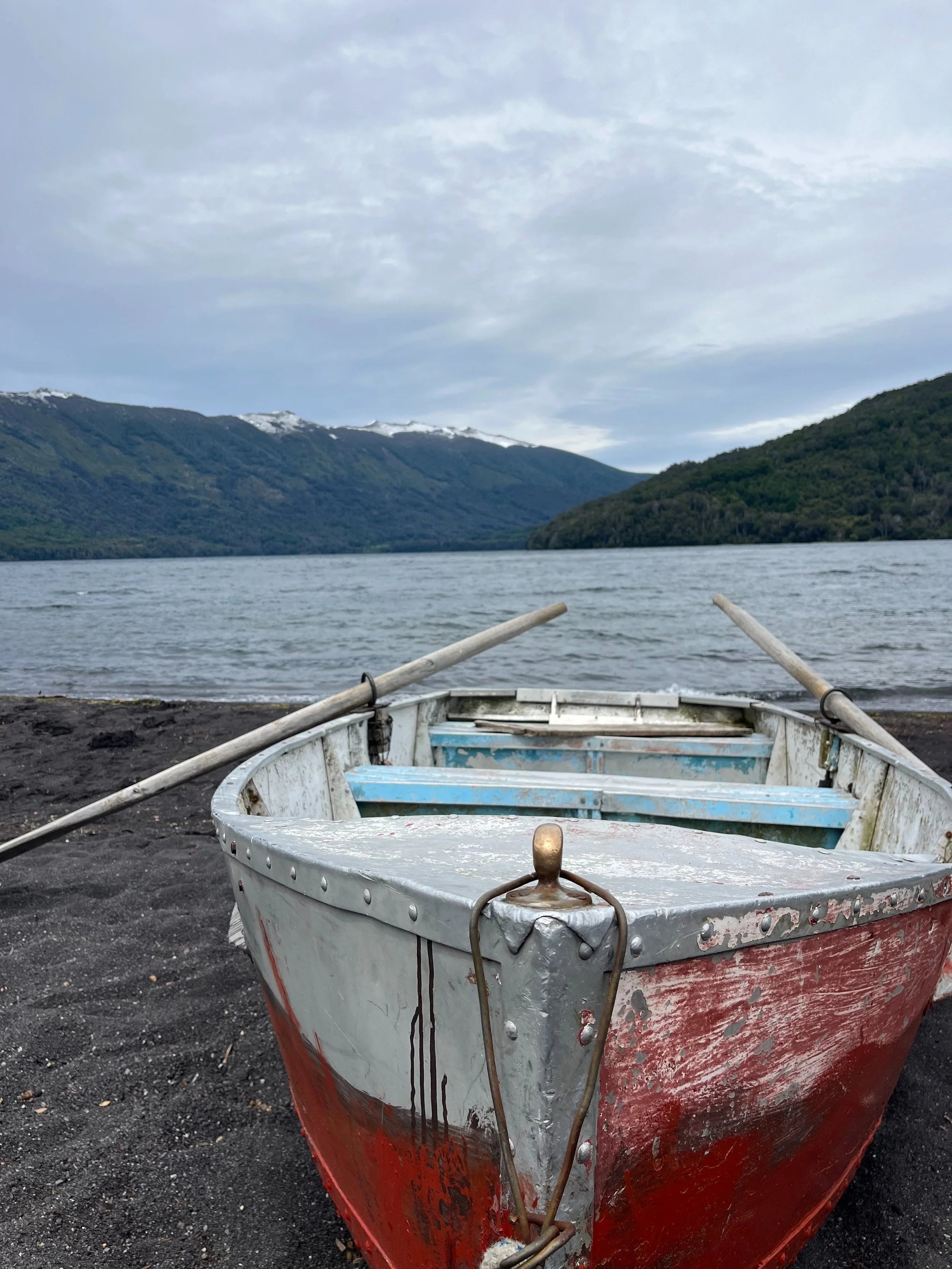 Old weathered boat resting on a dark sandy shore with oars, overlooking a lake with mountains in the background under a cloudy sky in the Futa Mawiza Biocultural Territory spanning Argentina and Chile.