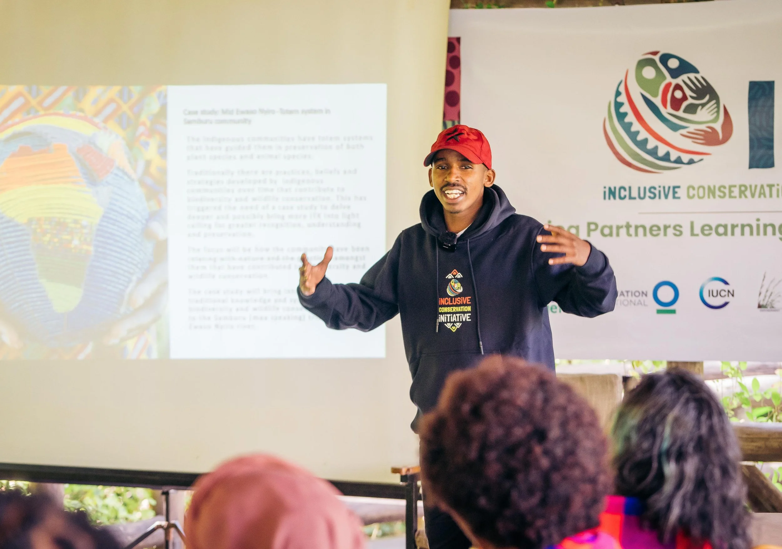 A man in a red cap and dark hoodie giving a presentation in front of a projector screen and a banner that reads 'Inclusive Conservation'. He is speaking to an audience with their backs visible in the foreground.
