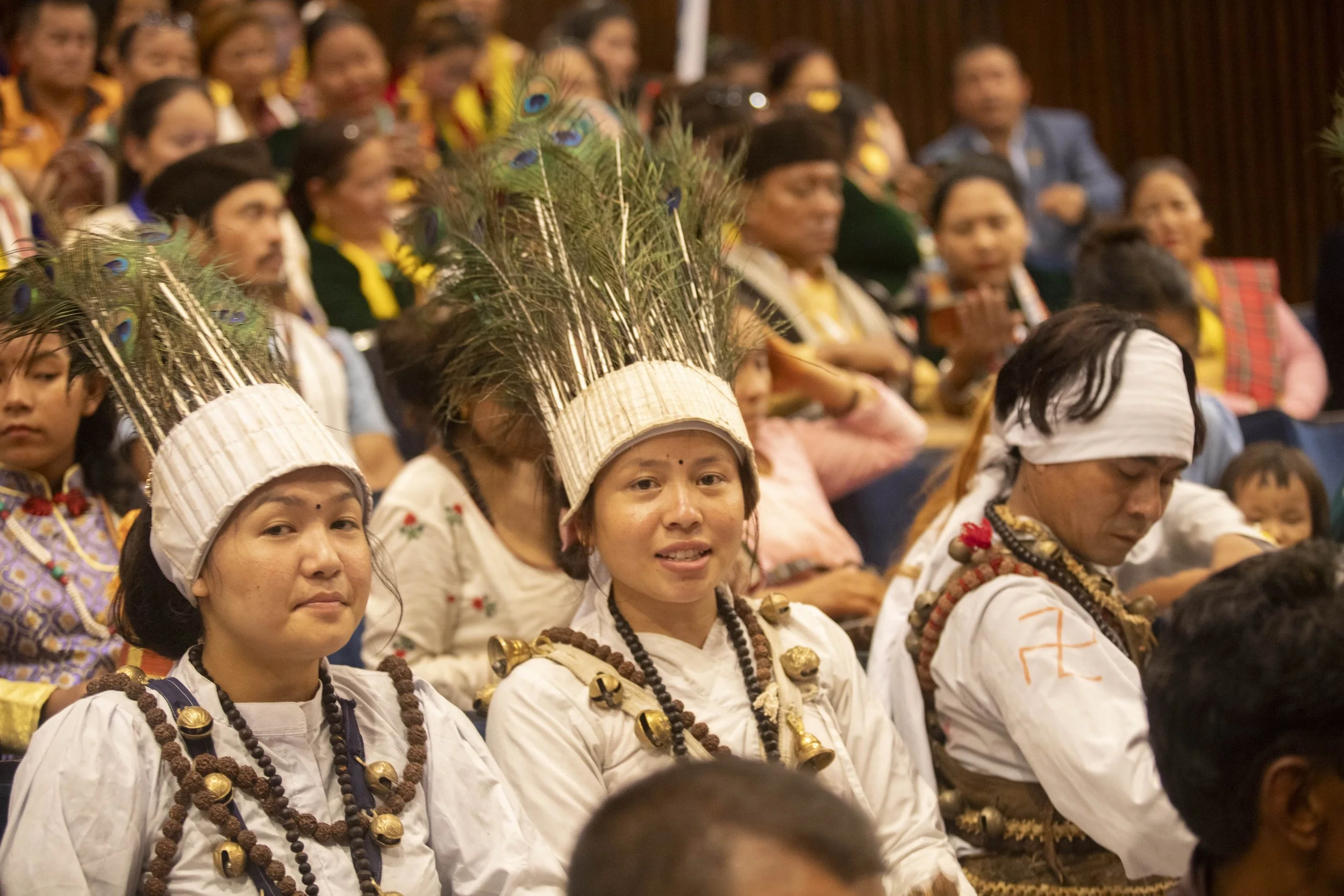 Indigenous Thai People attending a cultural event, with some wearing traditional attire and feathered headdresses, seated in an indoor hall.