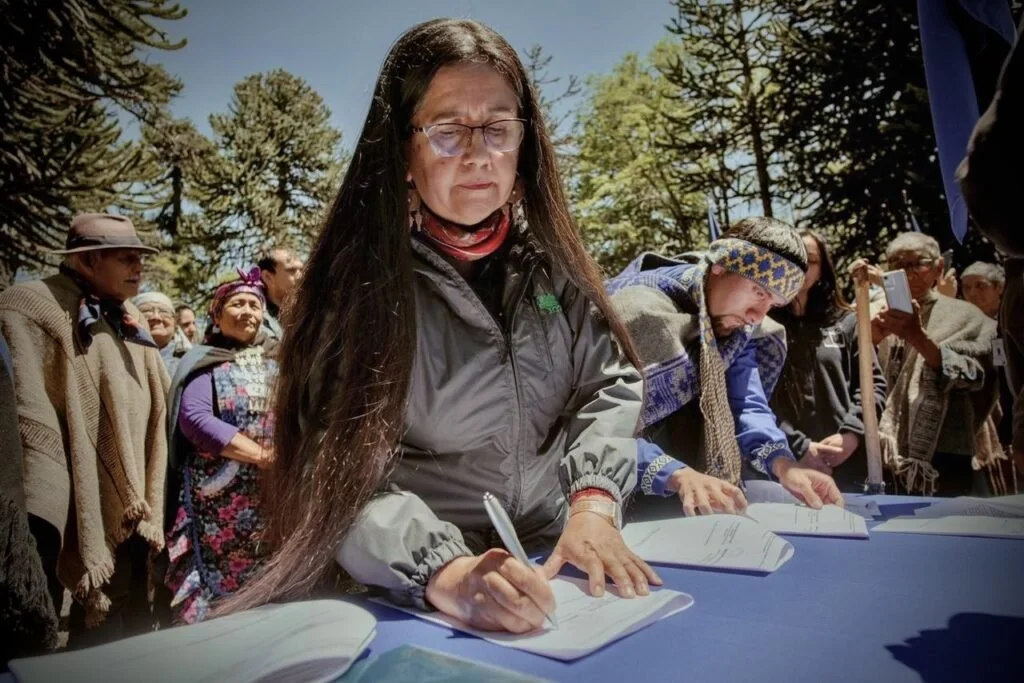 A woman with long dark hair, glasses, in a gray jacket, signing documents at an outdoor event, surrounded by people, trees, and clear blue sky in Chile.