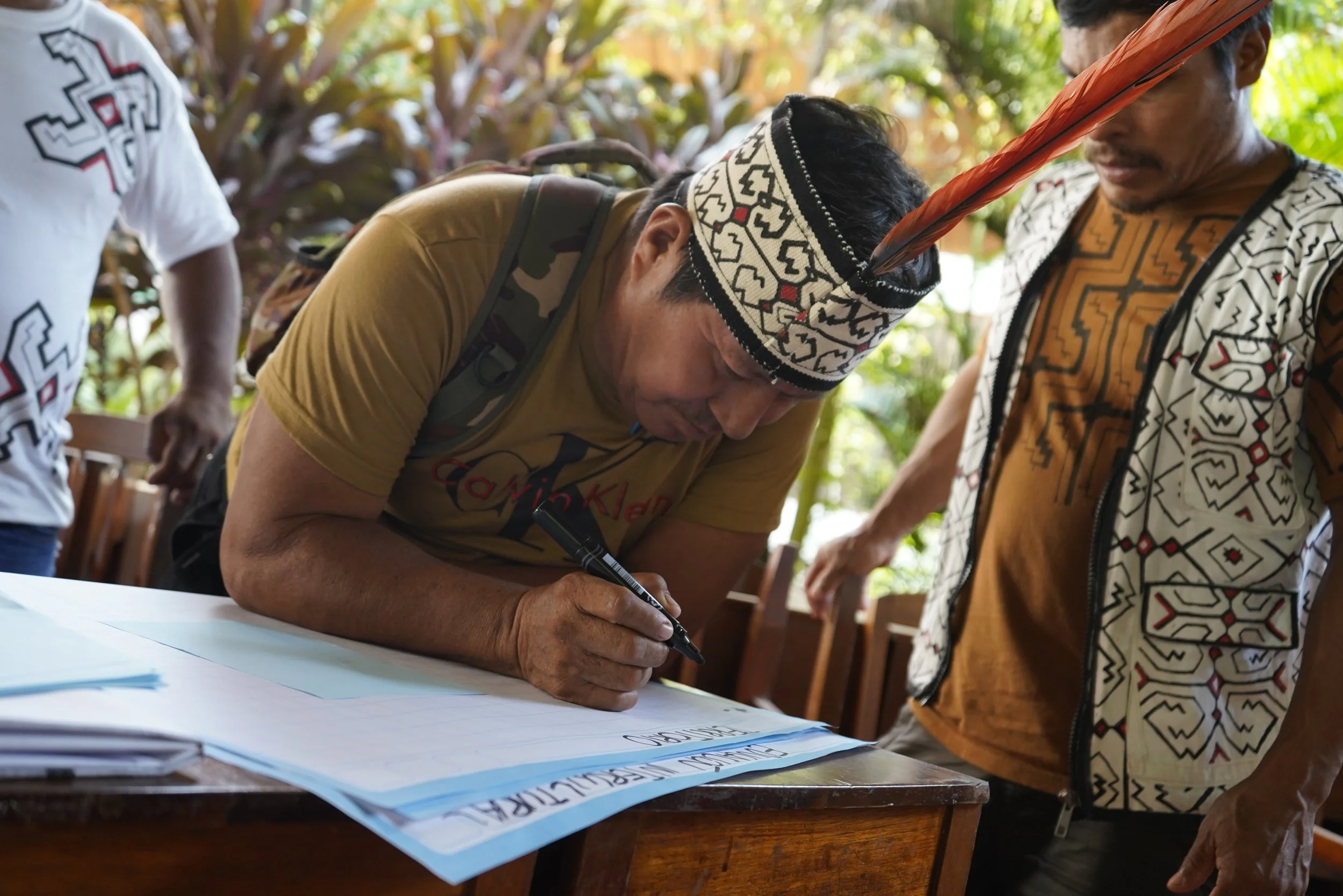 An Indigenous man from FENAMAD wearing a traditional Indigenous headband is signing documents on a table, with two other men standing nearby, outdoors with greenery in the background in Peru.