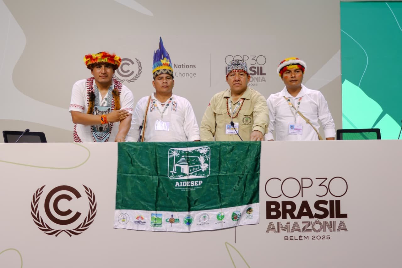 Four Indigenous men from the Southwest Amazon standing behind a table at COP30 Brazil Amazon, with a green flag and cultural accessories, during the conference in Belém 2025.