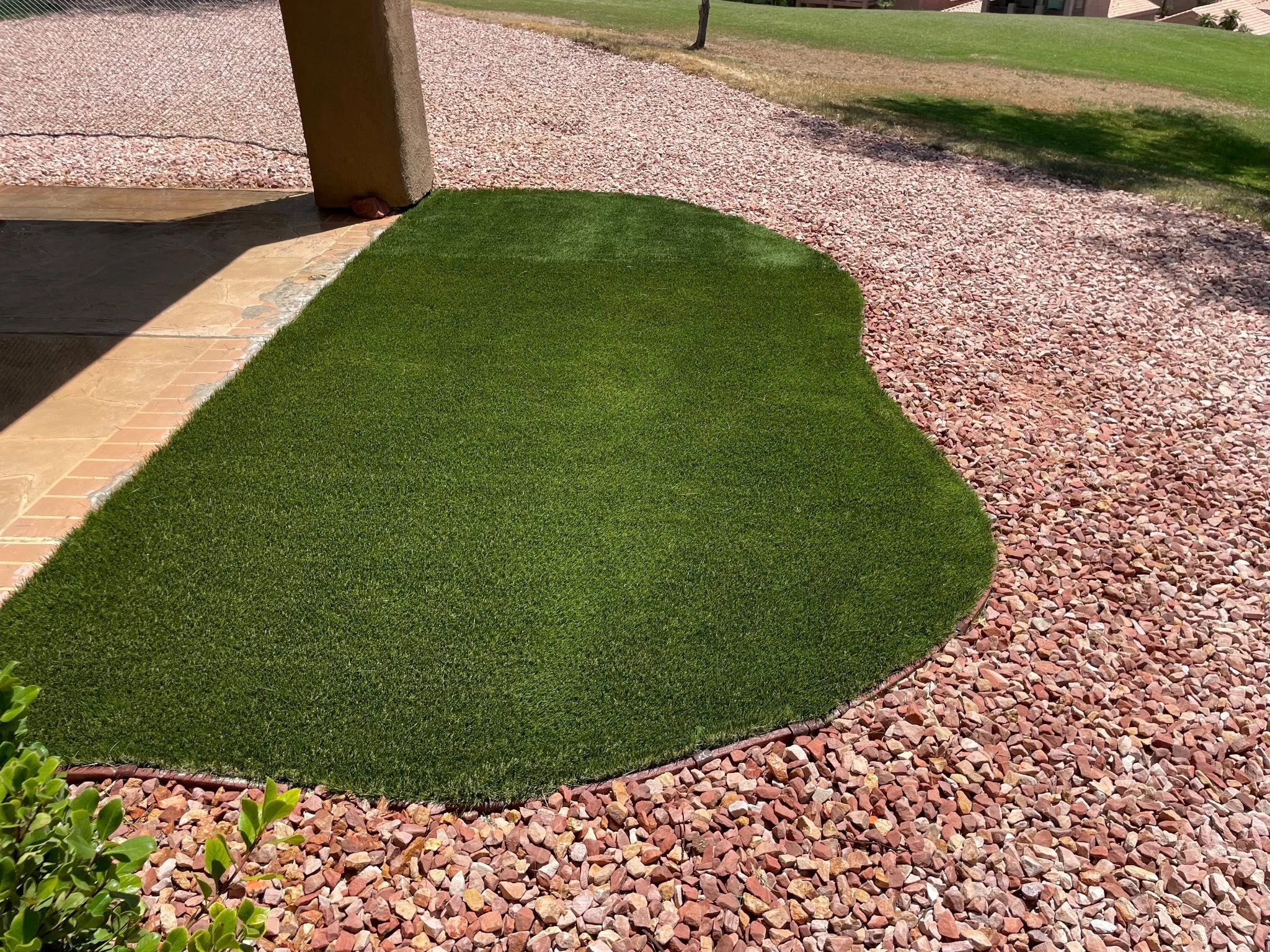 Close-up of a small, irregularly shaped patch of green artificial grass surrounded by reddish-brown gravel, next to a patio with stairs.