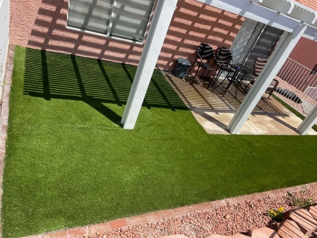 An outdoor patio with artificial grass, a small seating area with three chairs and a table, and a shadow pattern cast by a pergola. The patio is adjacent to a pink stucco building with a window, and a white railing surrounds the space.