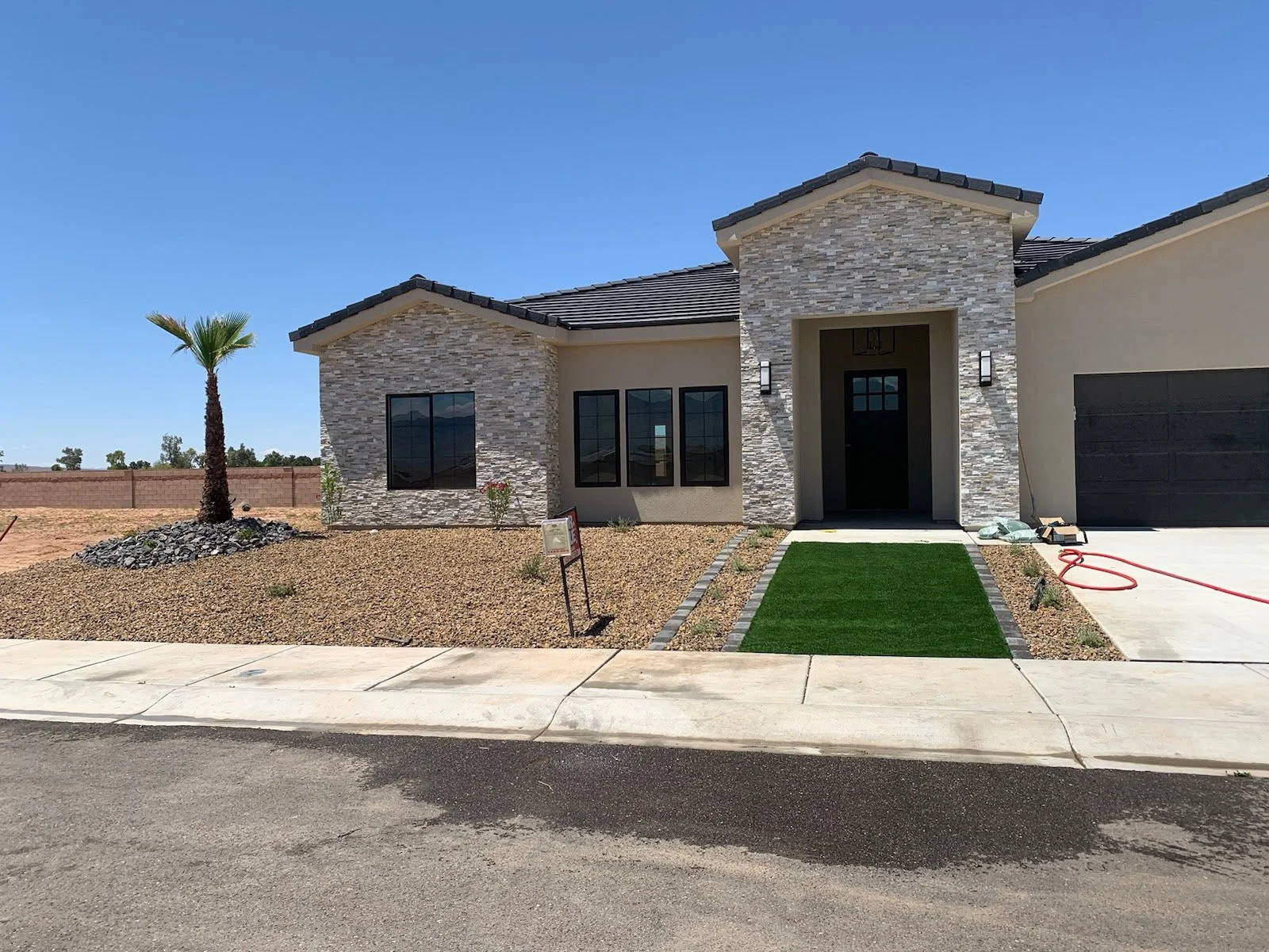 Front view of a new modern house with a small front lawn, a sidewalk, a palm tree, a stone and stucco exterior, and a black garage door. There is a red hose and some construction materials on the right side.