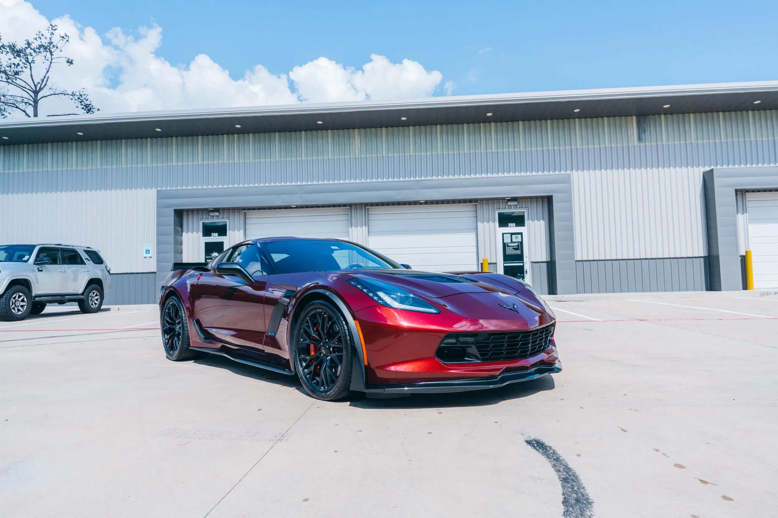 A red sports car parked in front of a commercial building with garage doors, with another white SUV nearby, under a partly cloudy sky.