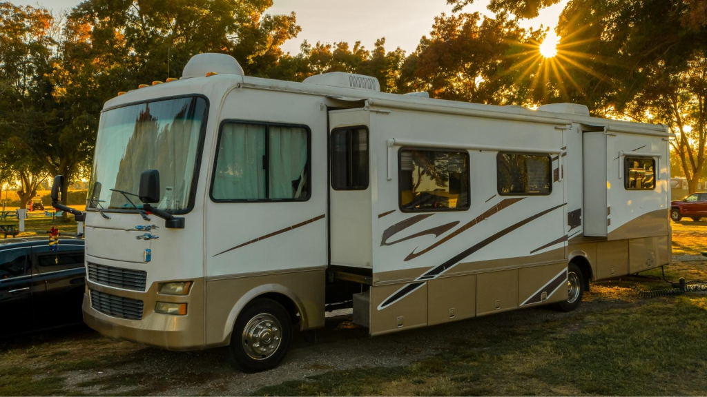A white and tan recreational vehicle parked on a grassy area during sunset with trees and a red car in the background.