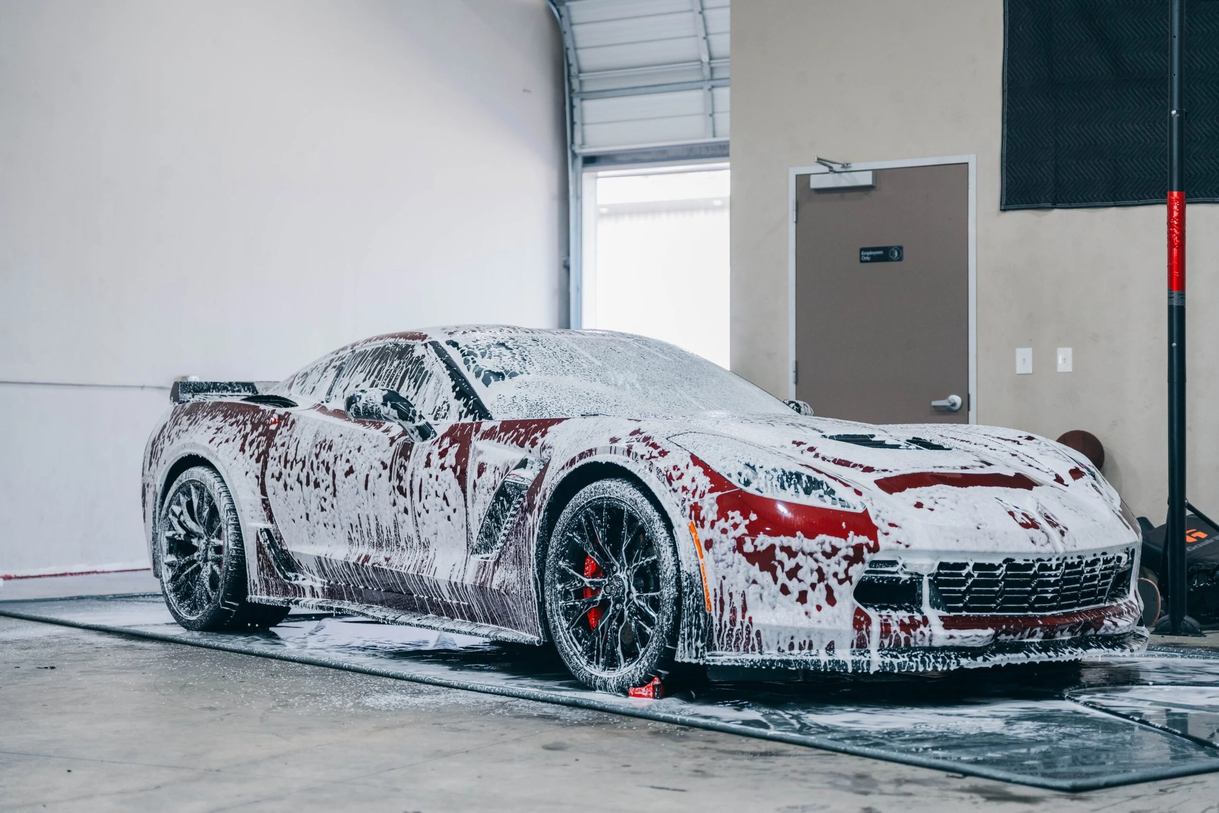 Red sports car covered in white soap suds during a car wash in a garage.