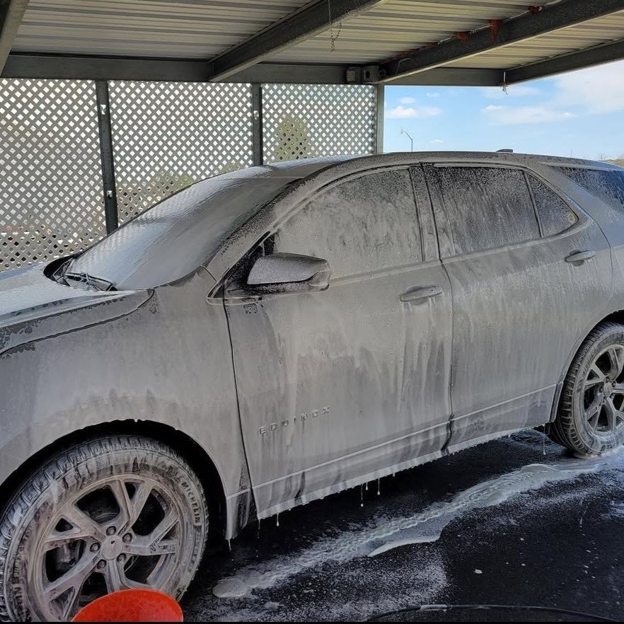 Dusty gray Honda sedan covered with soap foam, parked under a carport, with a red bucket in front.