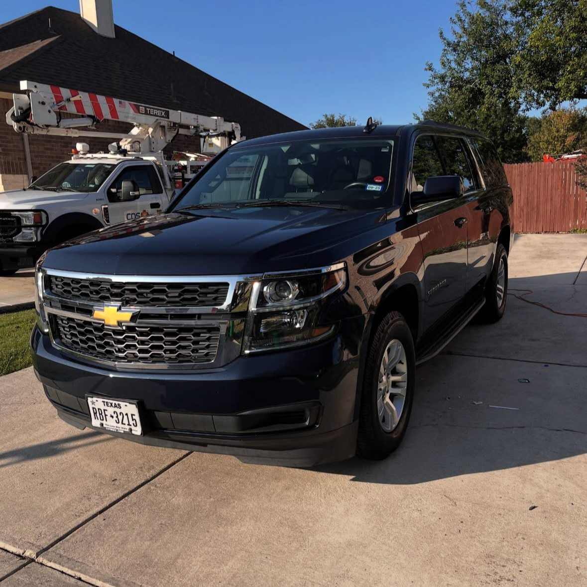 A black Chevrolet SUV parked on a driveway with a Texas license plate, next to a utility truck with a bucket lift in a residential area during daytime.