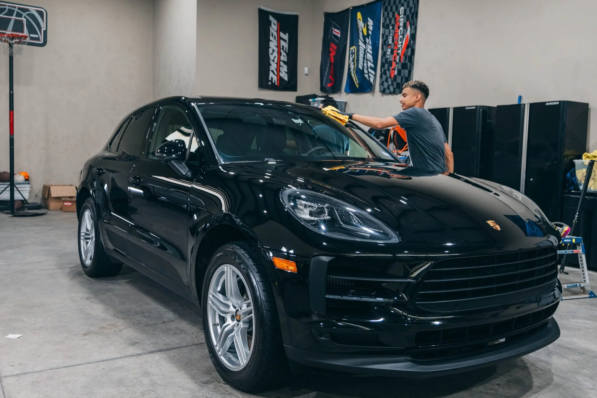 A young man cleaning a black Porsche SUV in a garage with banners on the wall and storage cabinets in the background.
