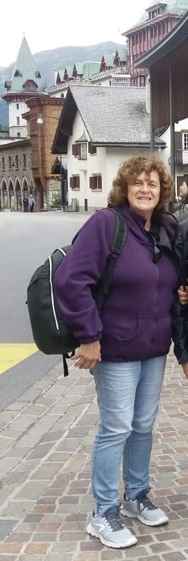 Donna sorridente con giacca viola e zaino, standing su una strada di pietra, con edifici storici e montagne sullo sfondo.