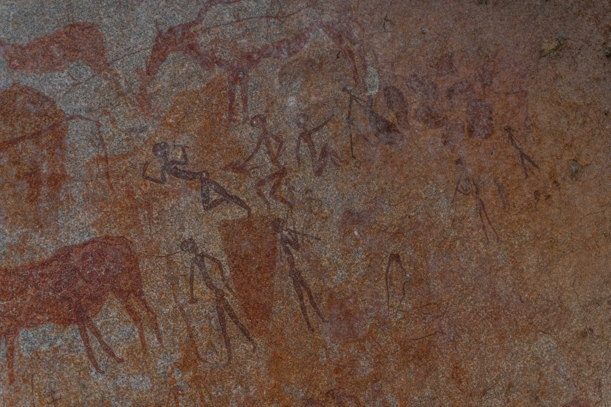 Ancient cave painting depicting human figures and animals on a reddish-brown rock surface.