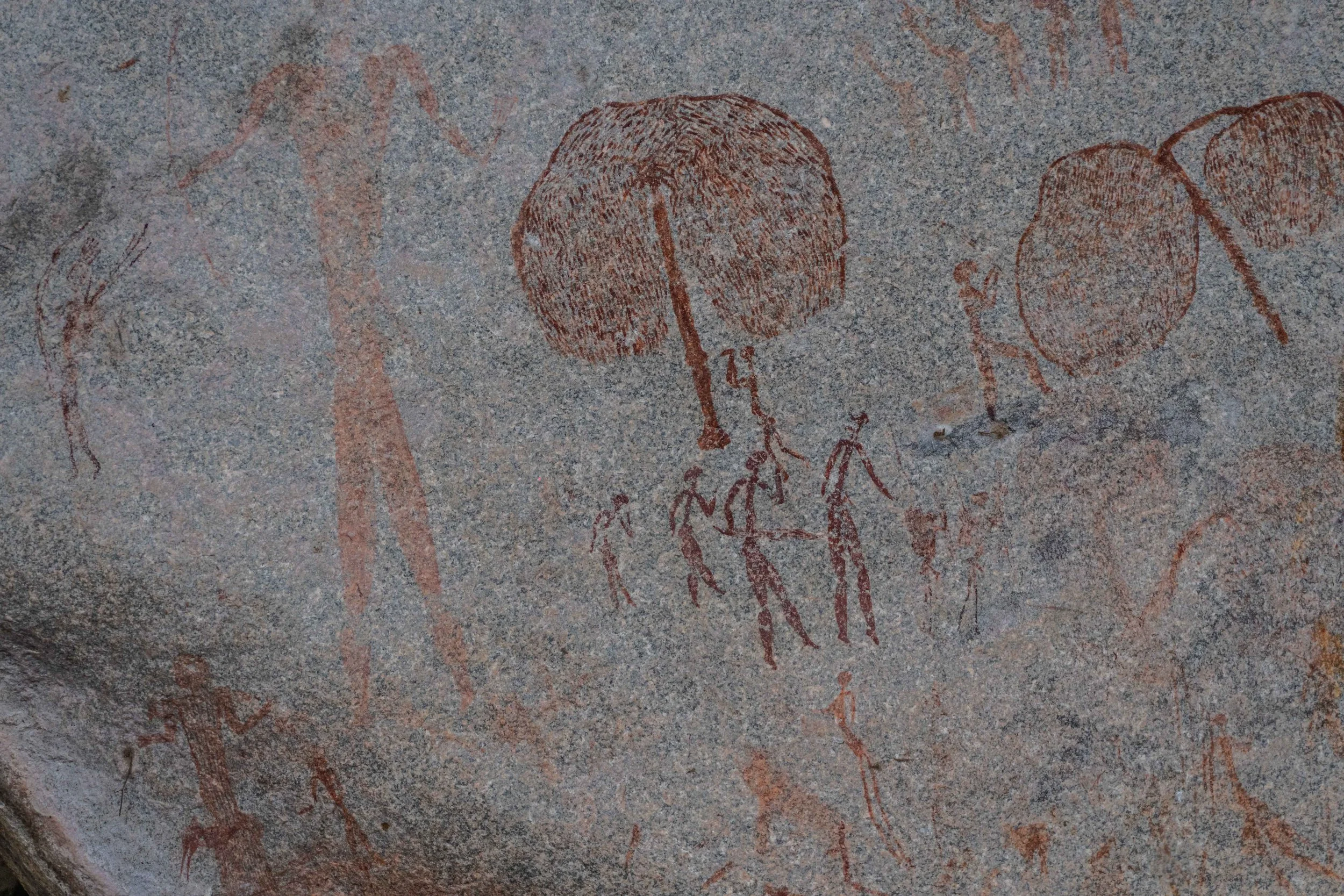 Prehistoric cave painting depicting dancing human figures, elongated human figures and tree depictions in red pigment.