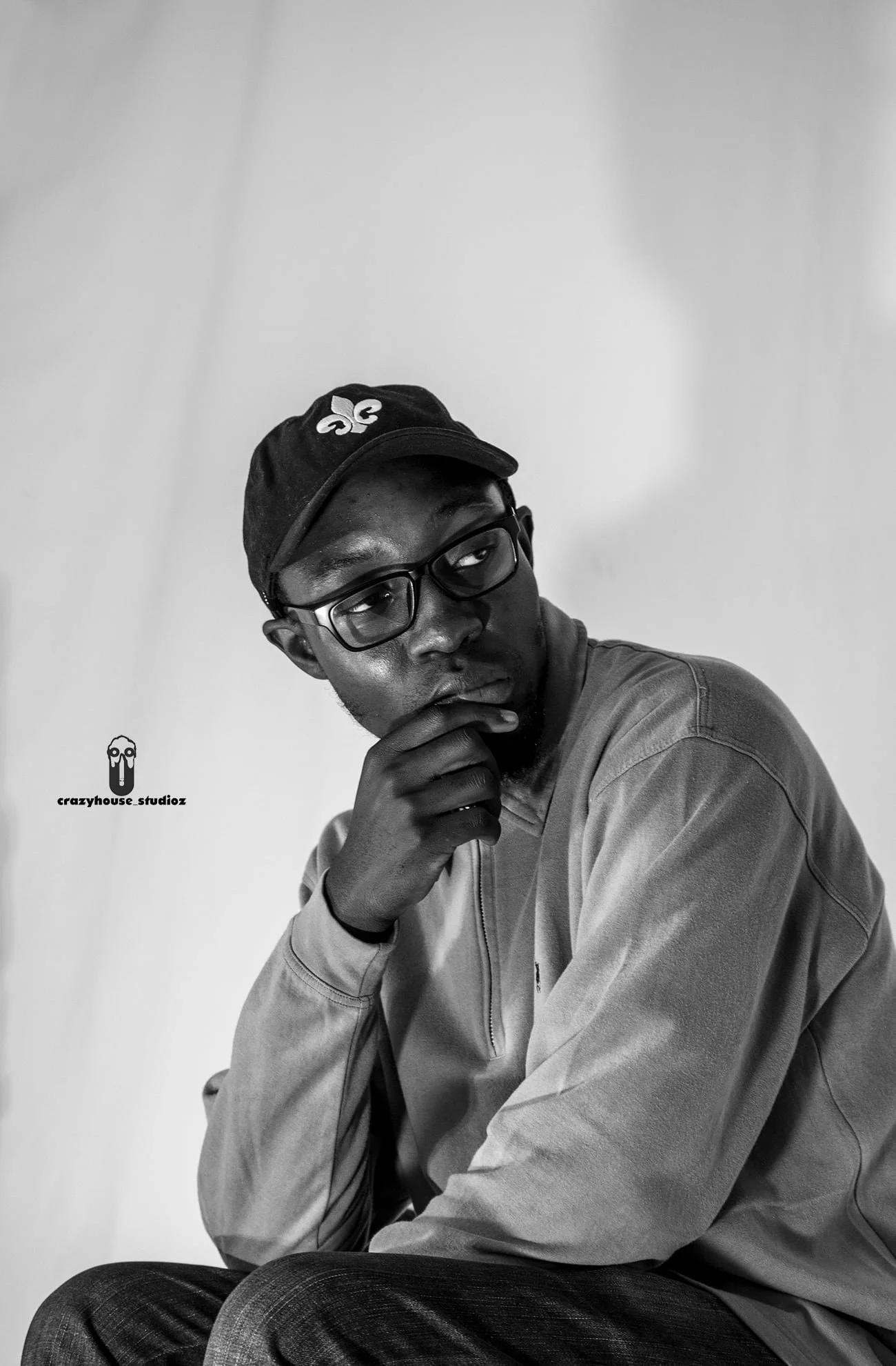 Black and white portrait of a young man (tawanda the photographer) wearing glasses, a cap, and a casual jacket, sitting with his hand on his chin against a plain background.