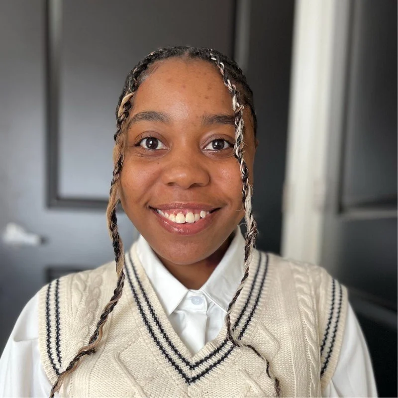 Young girl (the author, Schyler) with braided hair and a bright smile, wearing a white collared shirt and a cream-colored vest with black stripes.