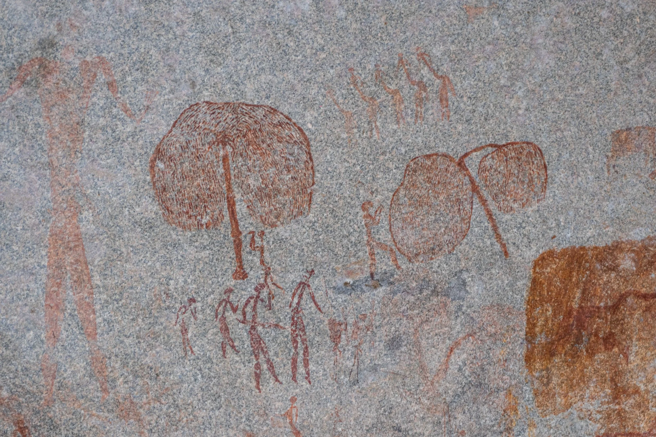 Ancient cave painting depicting humanoid figures and trees, in reddish-brown pigment.