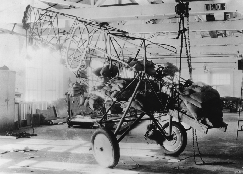 Black and white photo of an old, partially assembled aircraft in a hangar, with the frame and engine visible and covered with bags or padding, and a 'No Smoking' sign on the wall.