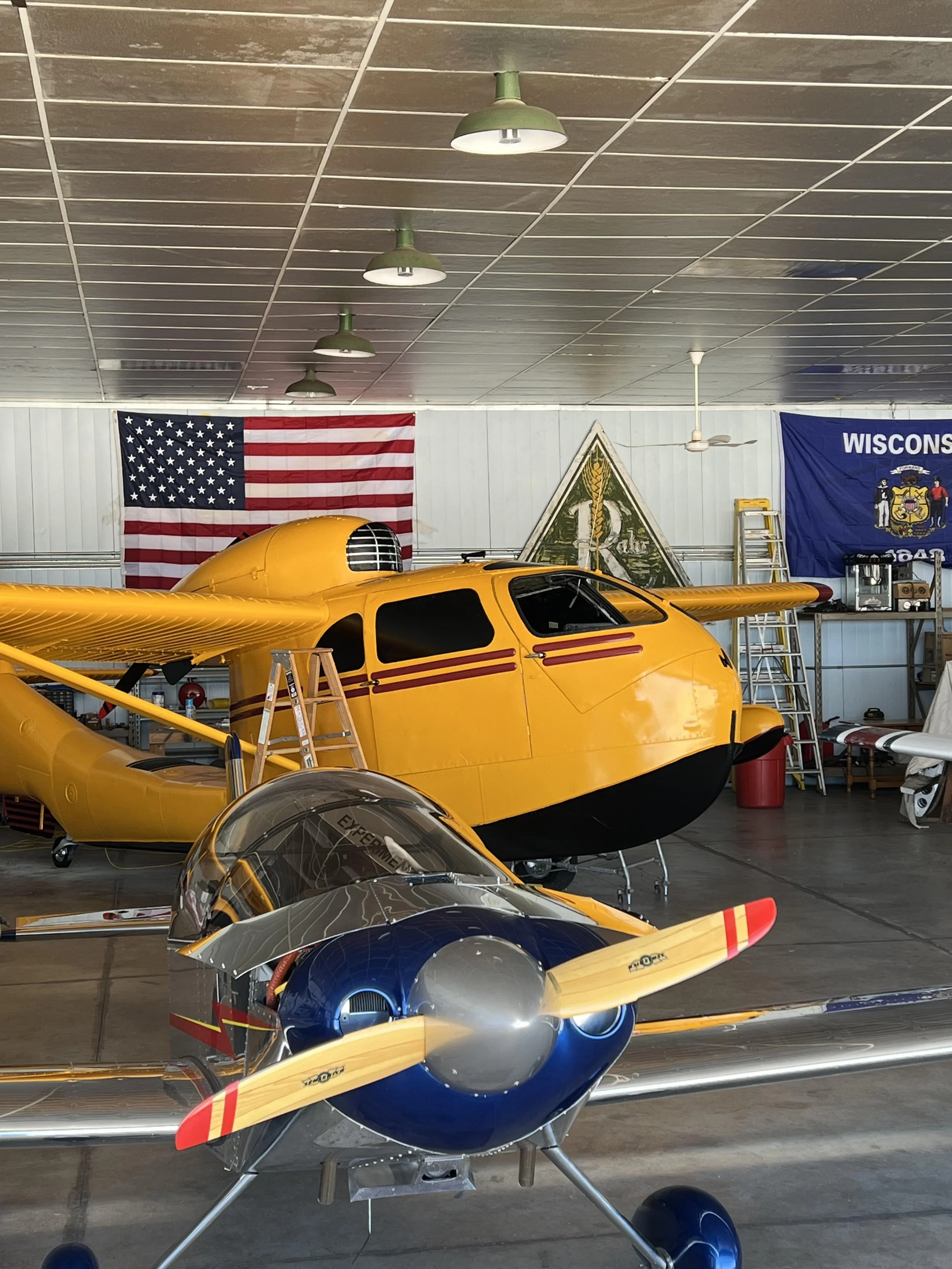 Indoor aviation museum with a small blue and silver airplane in the foreground and a yellow rescue aircraft with a glass-enclosed cockpit in the background. American flags and banners are displayed on the wall.