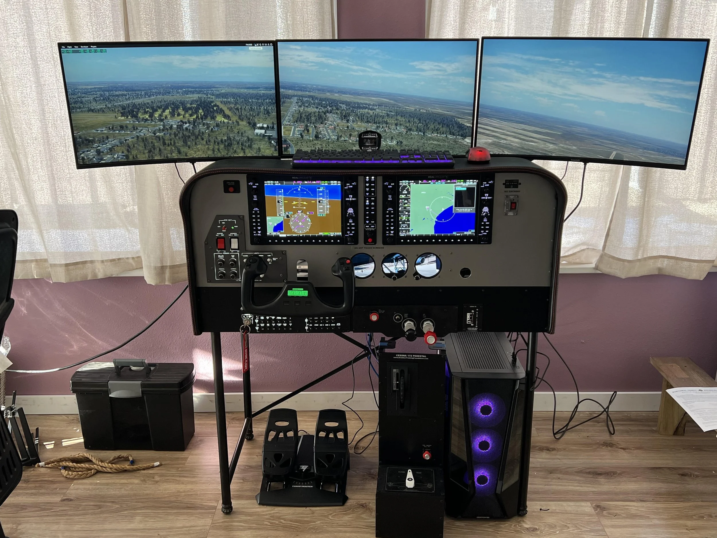 A flight simulator setup with three large monitors showing an aerial view outside a cockpit, a control panel with screens and controls, a keyboard and mouse, and a computer tower with purple lighting.