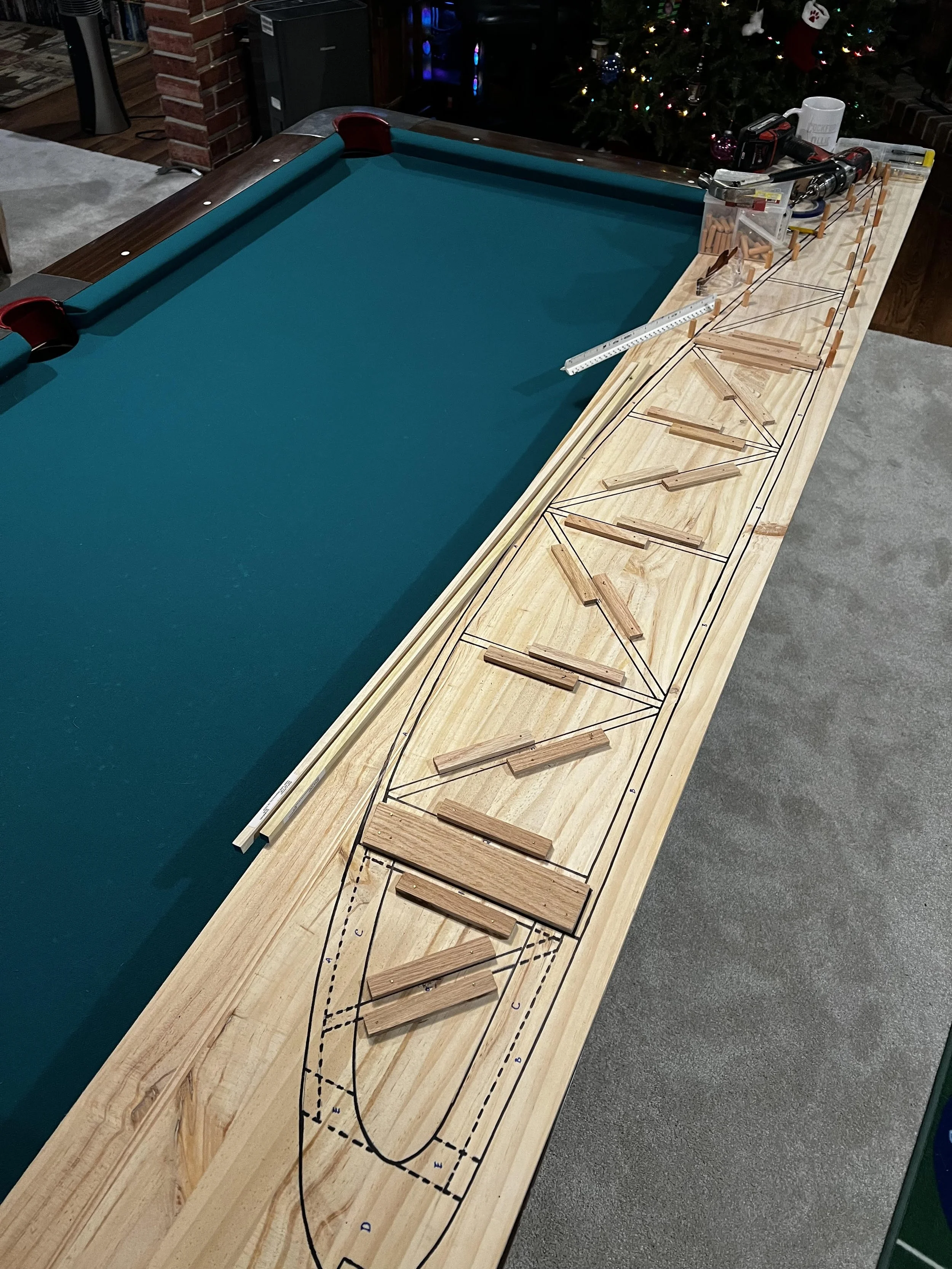 A pool table is being built, with a wooden frame on a workbench. The frame has a detailed blueprint drawn on it, and various wooden parts and tools are placed on top. In the background, there's a Christmas tree decorated with lights and ornaments.
