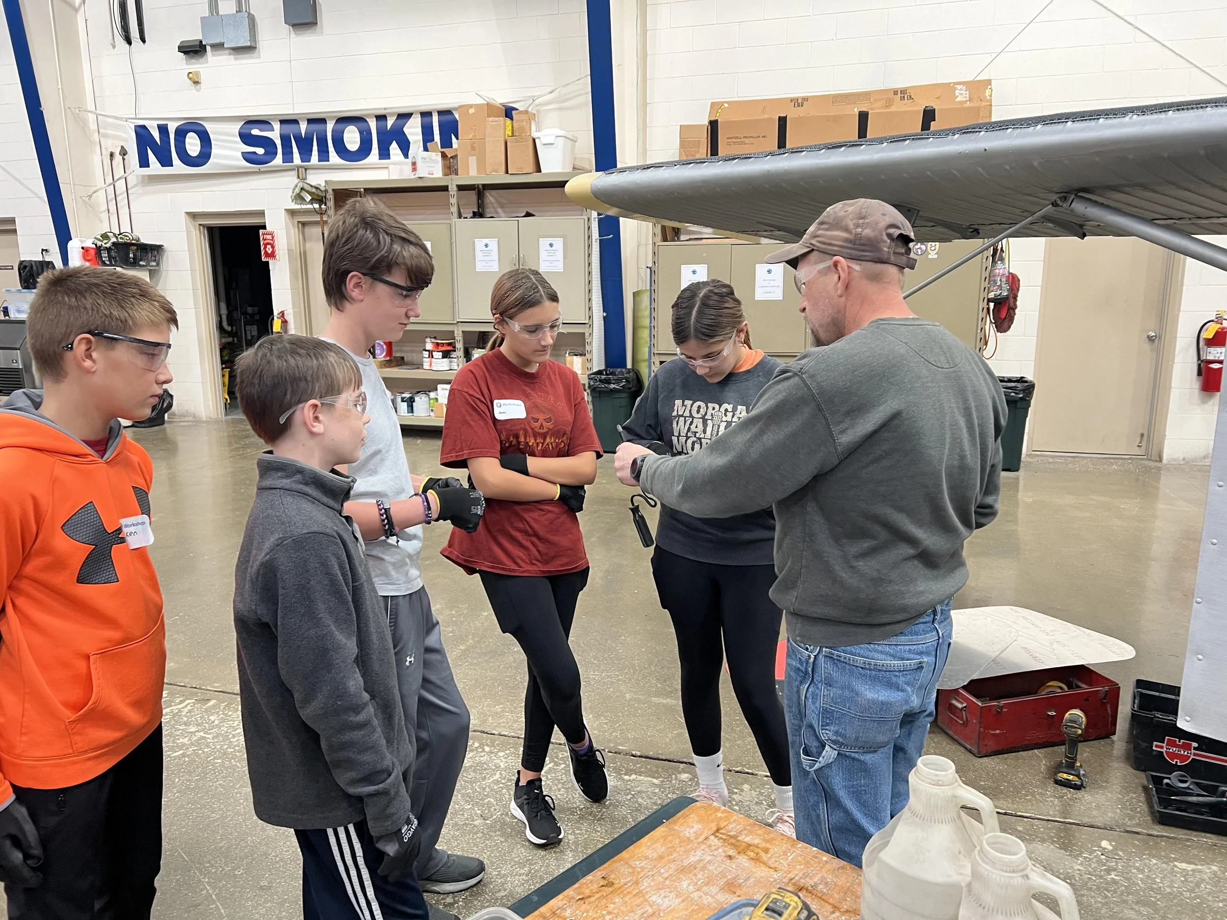 An instructor demonstrating something to five children in a workshop of an indoor facility, with the children attentive and some wearing safety glasses.