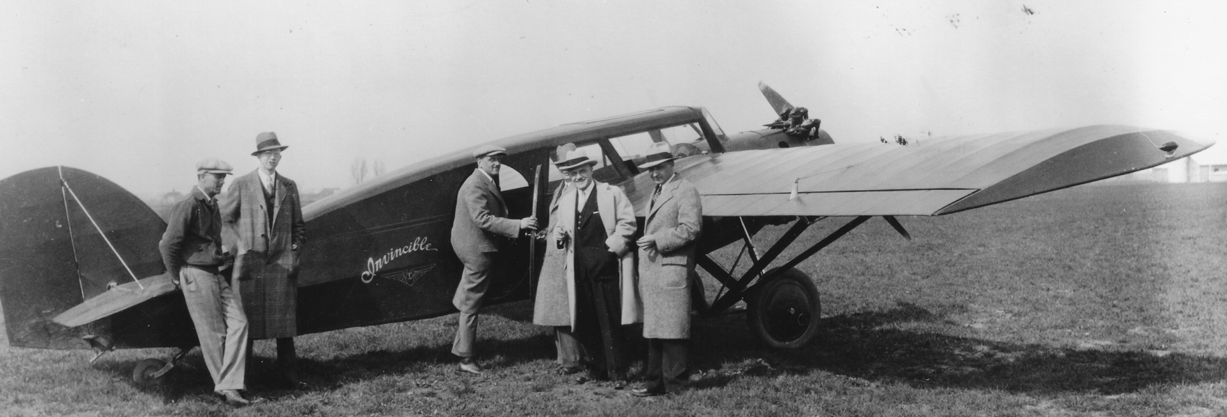 Black and white photo of five men standing next to a vintage airplane on a grassy field, with a tent in the background.