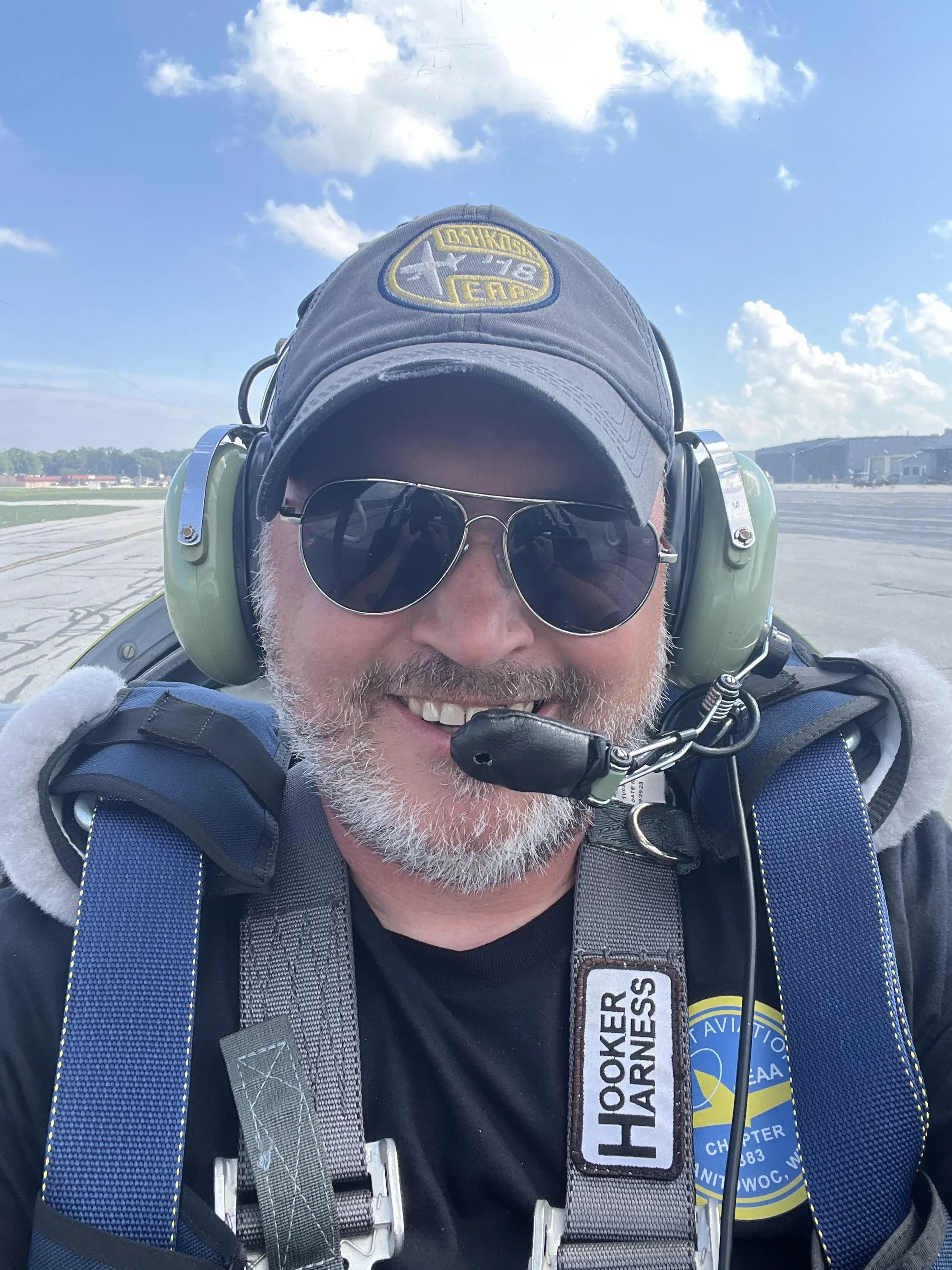 Smiling man in aviator sunglasses and pilot gear, including headset and cap, standing in front of an airport runway on a clear day.