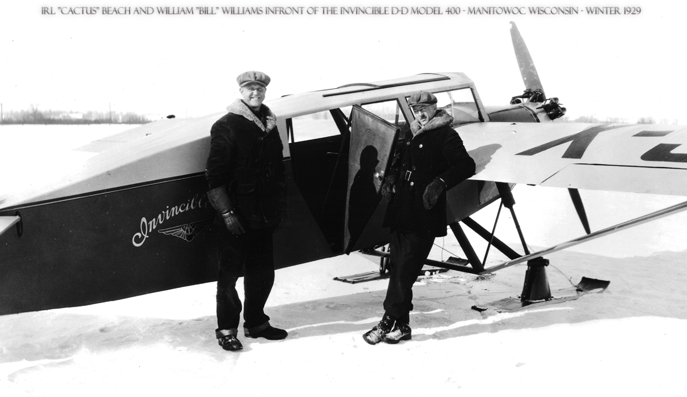 Two men in winter clothing and hats standing next to a vintage airplane on snow, with one man inside the plane and the other outside. The plane has the word 'Aviacil' on the side and is parked on snowy ground, with a flat, open landscape in the background.