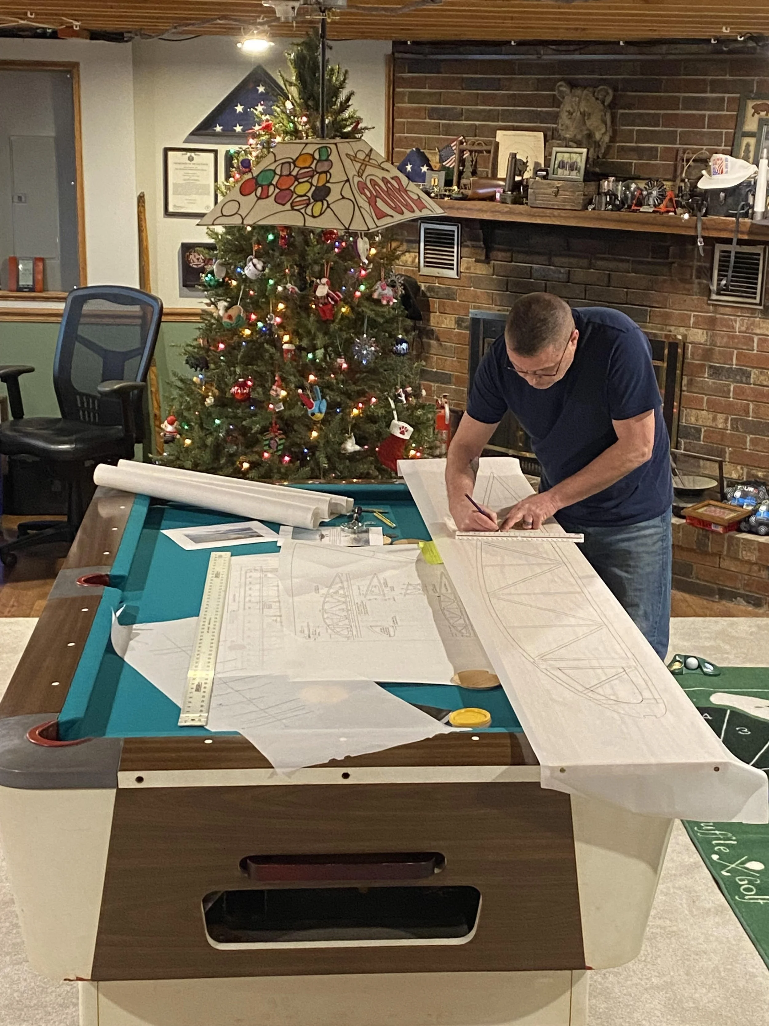 A man working on blueprints or architectural plans at a worktable with drafting tools, in a room decorated for Christmas with a decorated Christmas tree, a pie-shaped flag, and other seasonal decorations.