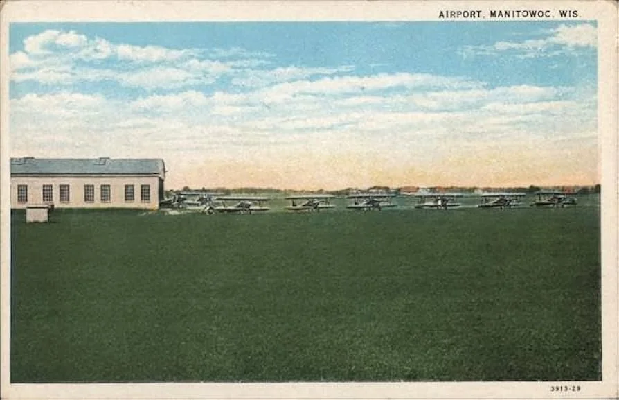 Historical photograph of Manitowoc, Wisconsin airport with small airplanes parked on the grass near a single large hangar building, under a partly cloudy sky.