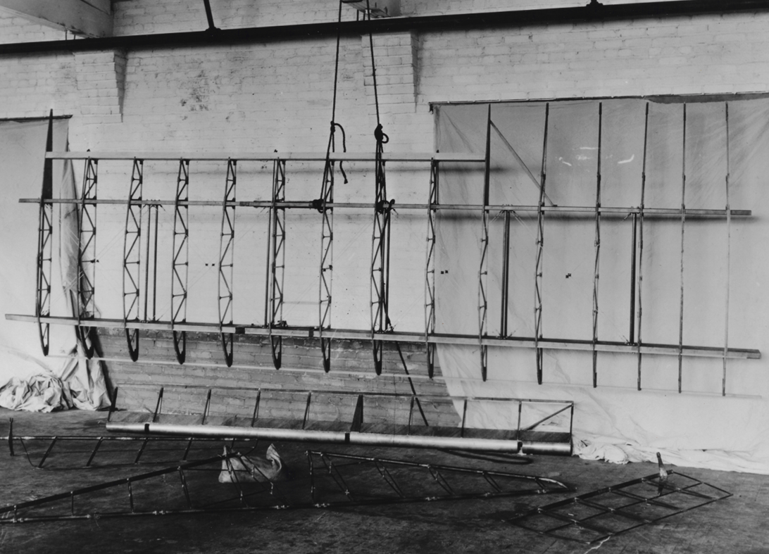 Black and white photo of a large metal aviation or drone wing frame, suspended in a workshop with various tools and materials around.