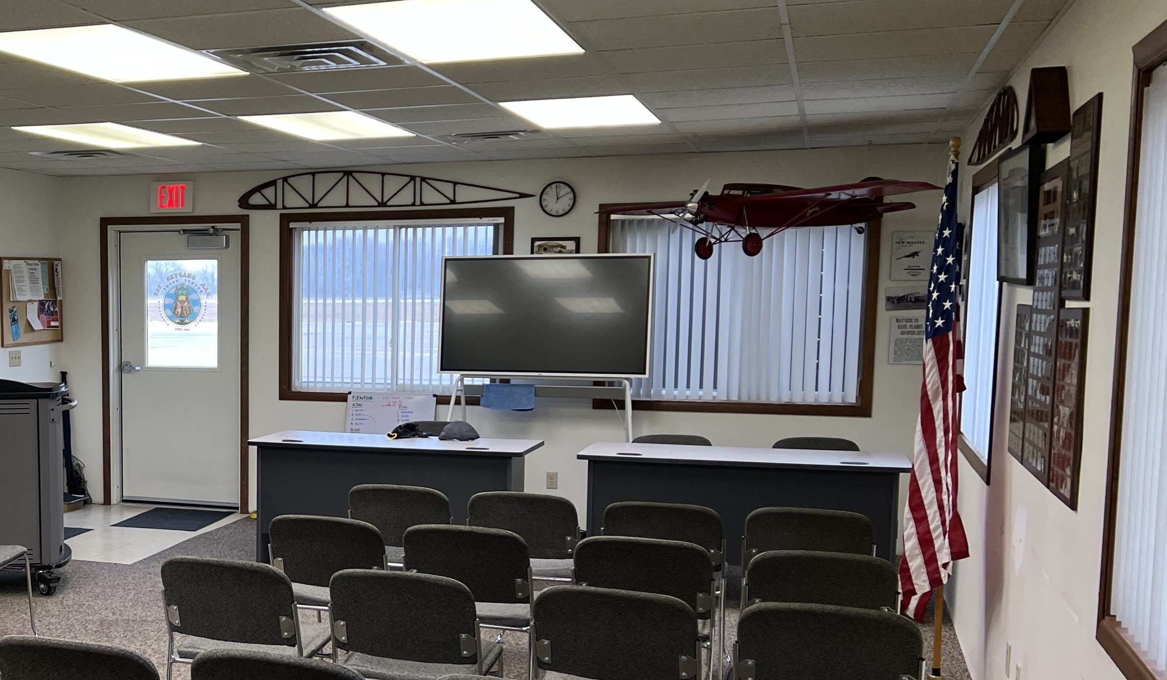 View of a small meeting room or classroom with chairs facing a large television on a stand, a flag, aircraft models, and various framed pictures and posters on the walls.