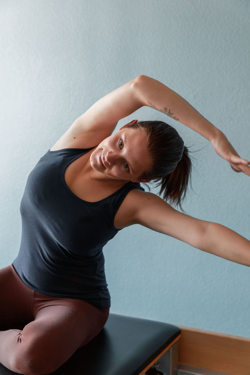 Woman doing a stretching exercise on a bench, wearing a black sleeveless top and maroon pants.