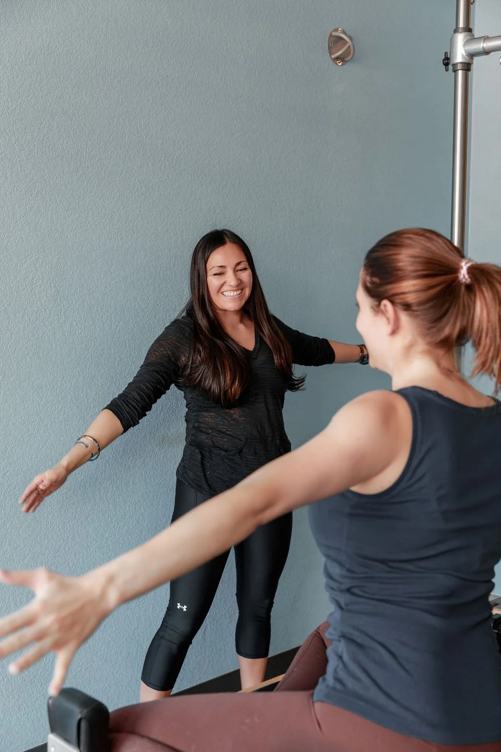 Two women in workout clothes smiling and stretching on exercise equipment in a fitness studio with blue walls.