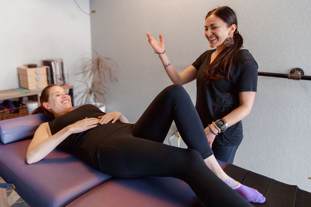 A woman lying on a treatment table smiling, while another woman, her instructor, laughs and gestures with her hand in a Pilates studio.