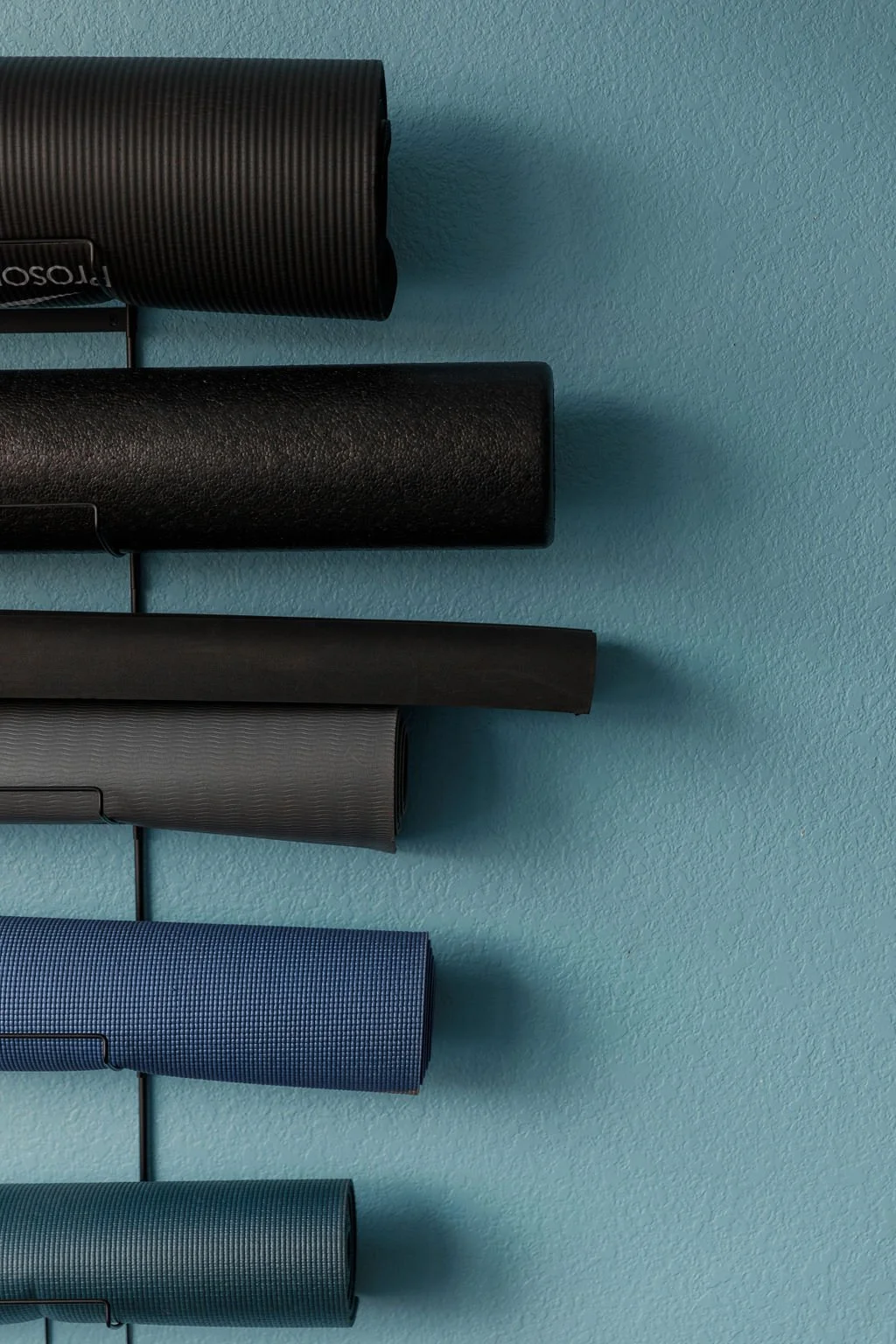 Four rolled-up yoga mats in black, dark gray, light gray, and blue, mounted on a wall with a light blue background.